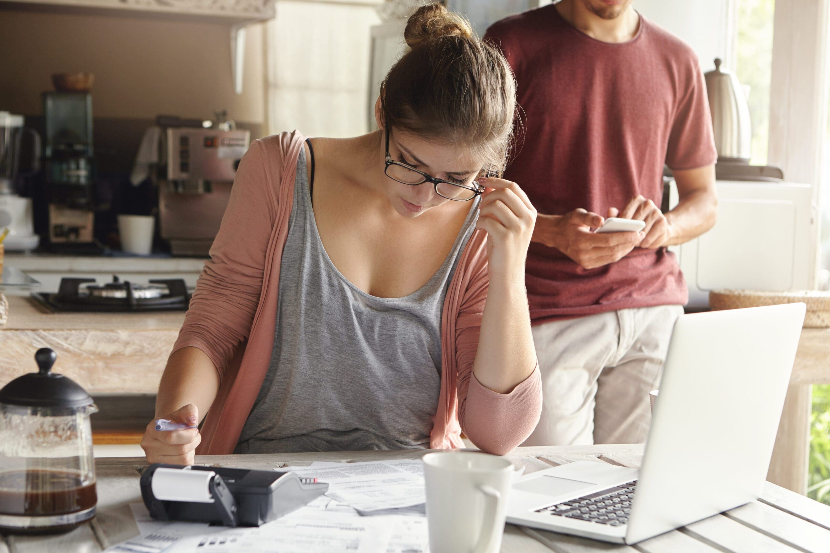 Woman in glasses working with papers and laptop, man in background.