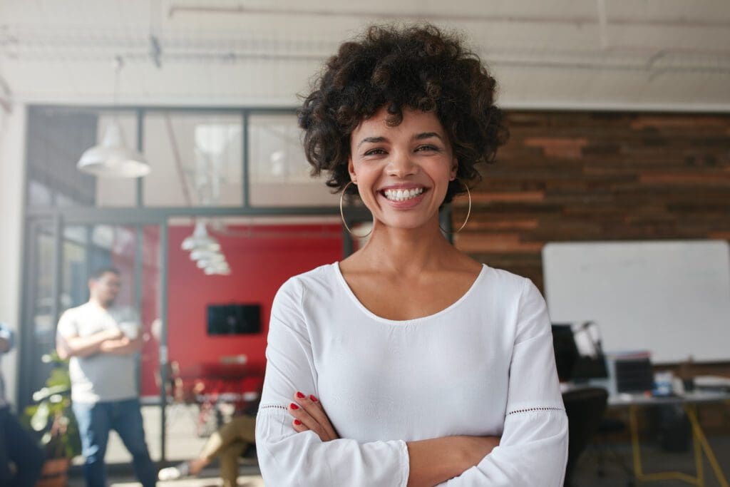 Smiling woman with curly hair and arms crossed, standing indoors.