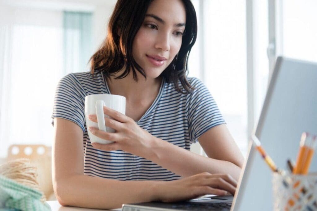 Young woman holding a cup while using a laptop.