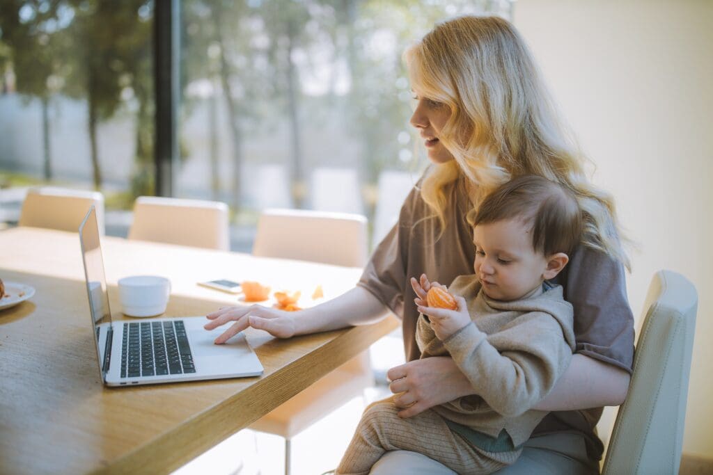 A woman working on a laptop while holding a baby.